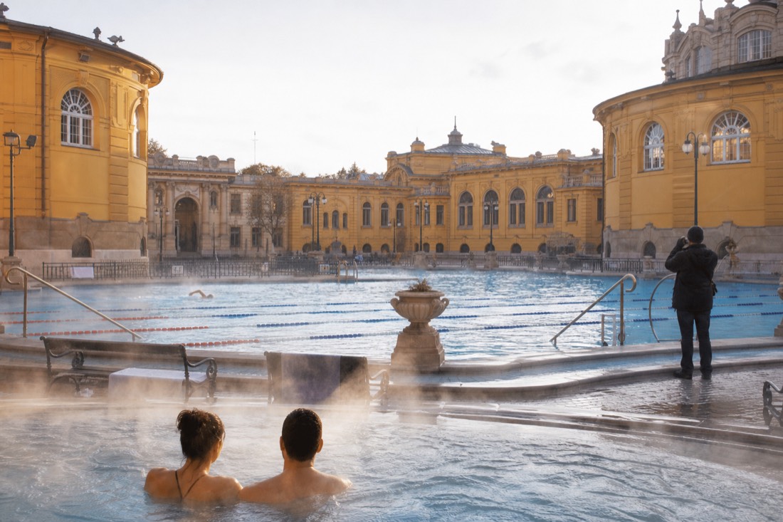 Different visitors at a Budapest thermal bath, a couple in the steaming pool, a swimmer in the lap pool and a photographer at the courtyard edge