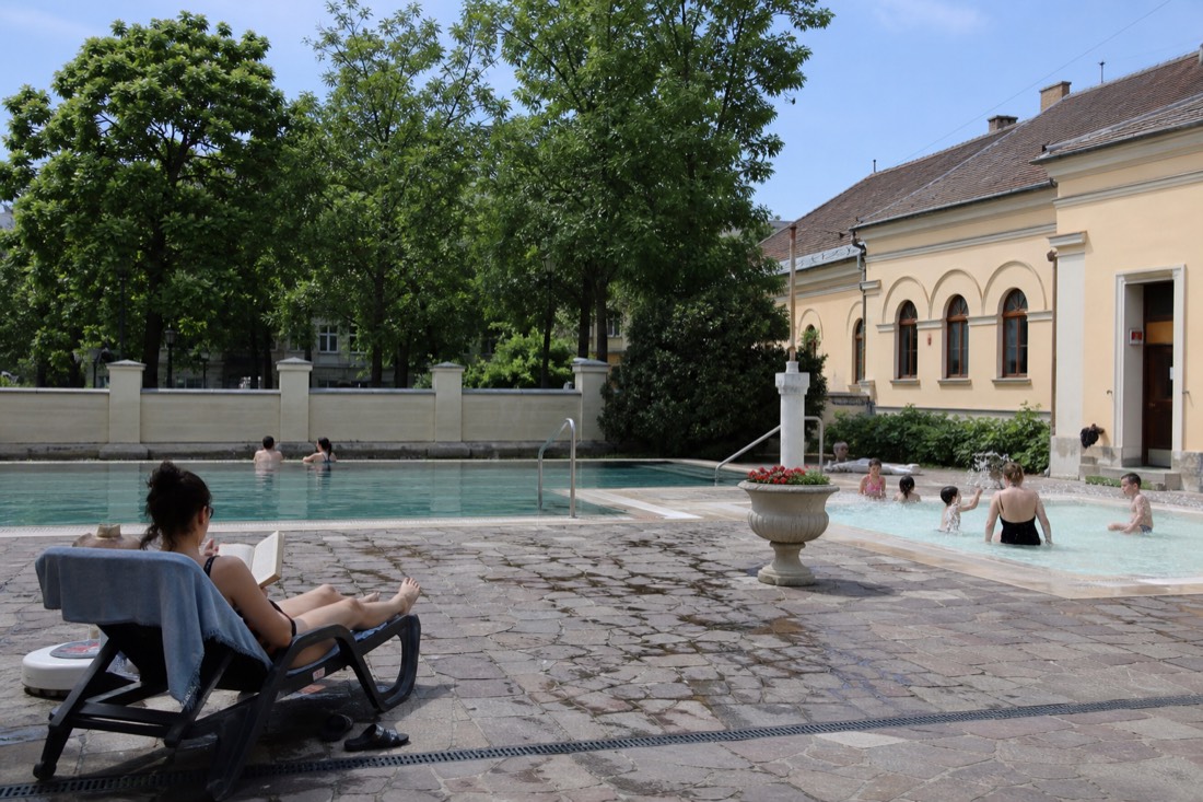 Different bathers, a solo reader, a couple and a family, spread across a calm Budapest bath complex