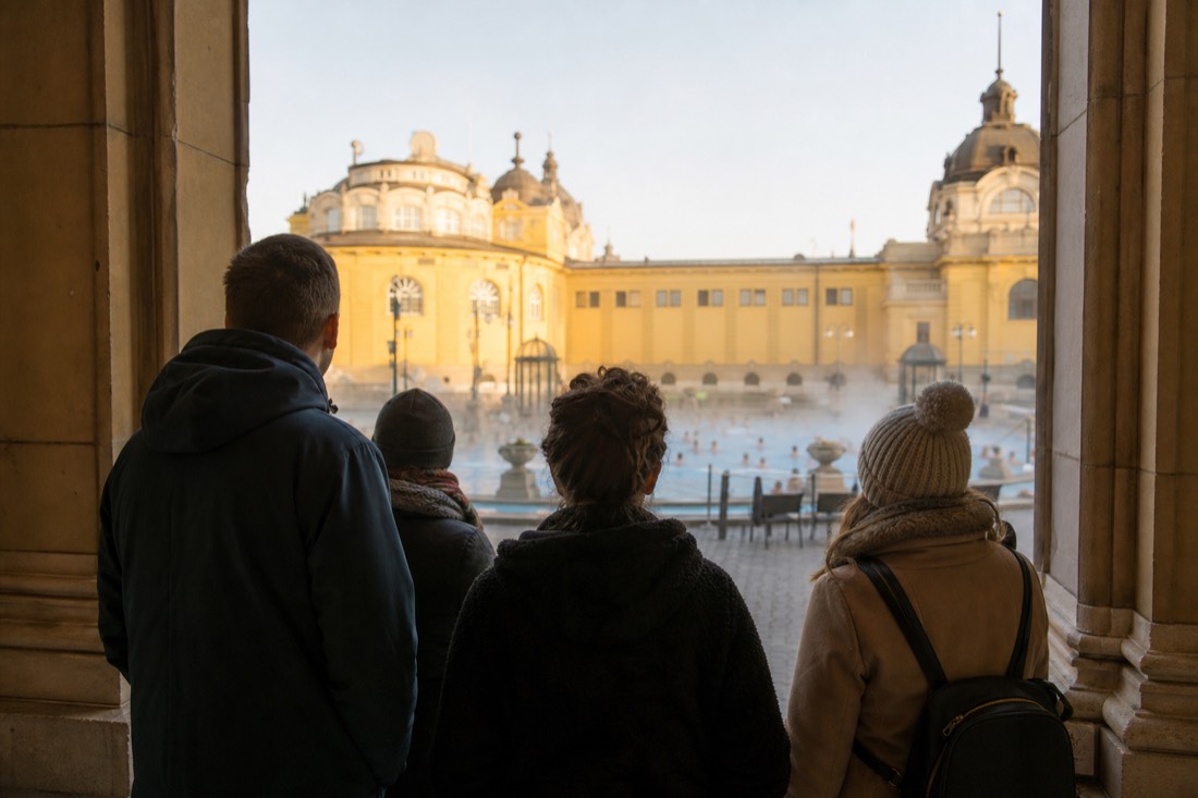 Small group of visitors seen from behind pausing at a bath palace courtyard entrance as steam rises off the pools beyond
