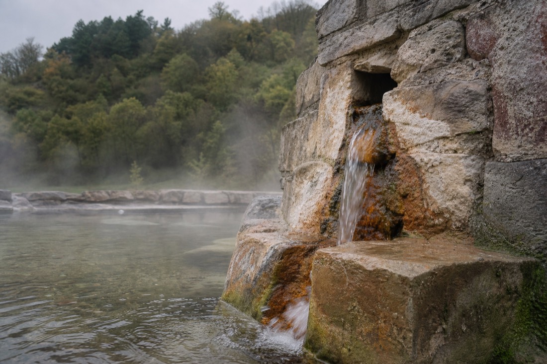 Weathered stone spring outlet with mineral staining at the foot of the Buda hills, water trickling into a small warm pool