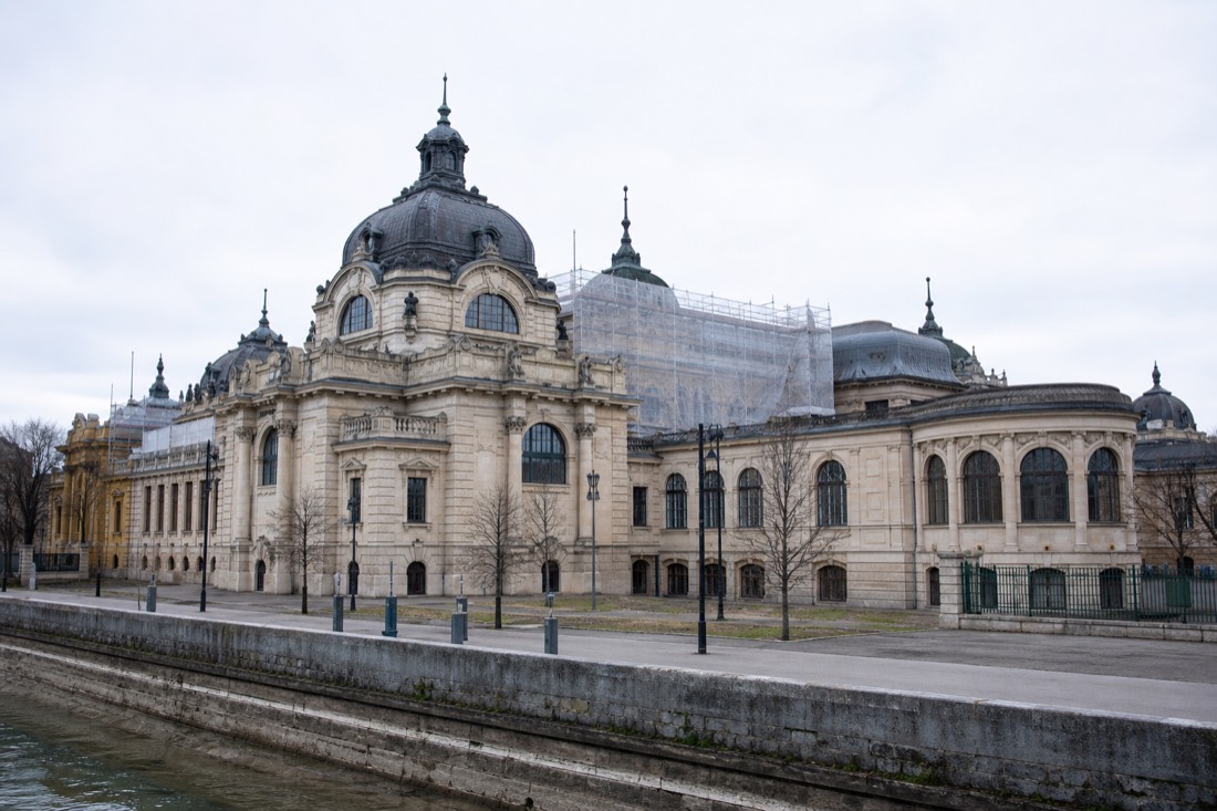 Historic bath building on the Buda embankment partly wrapped in renovation netting under a soft grey sky