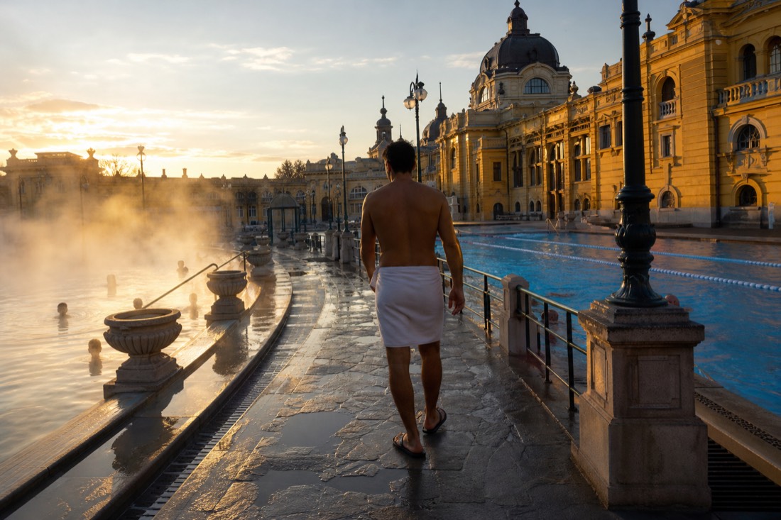 Bather walking along a wet stone poolside path between the outdoor pools of a Budapest thermal bath in early light