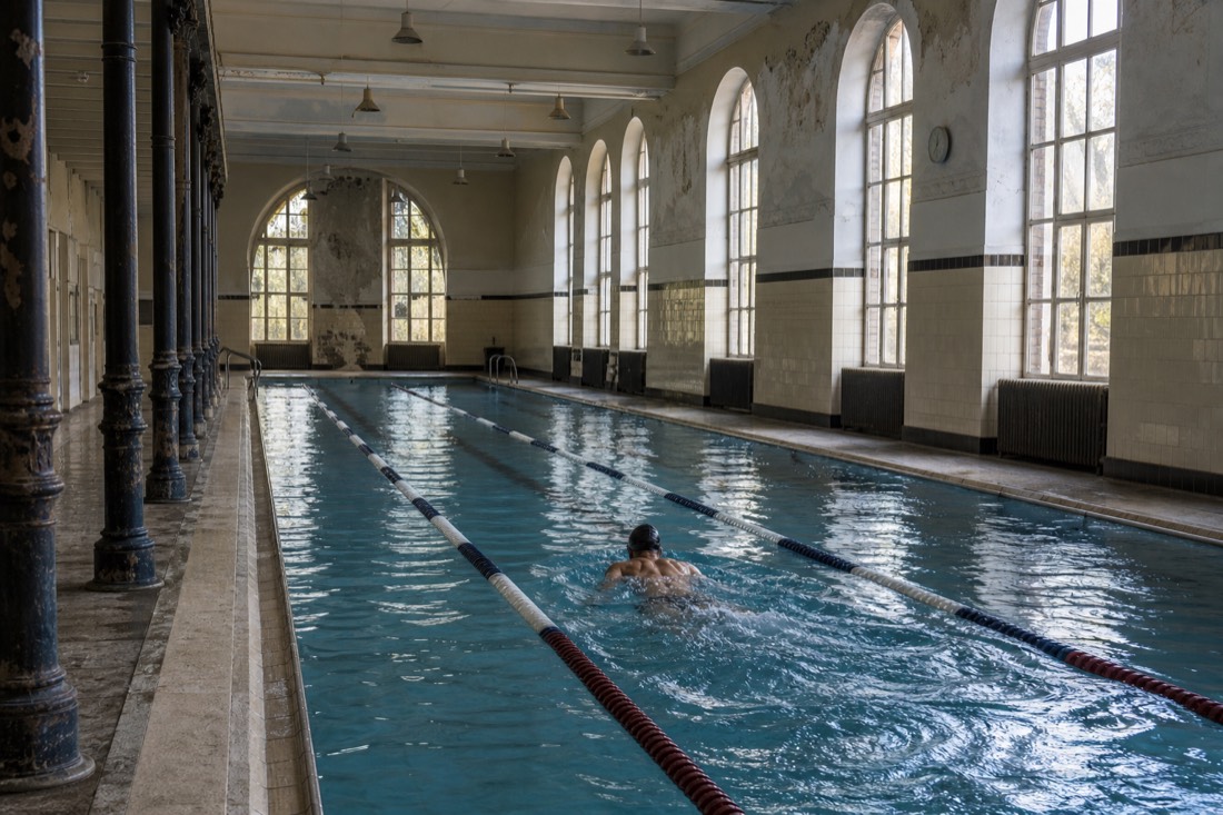 Indoor lane pool at a local Budapest thermal bath with a single early swimmer mid-length under high windows
