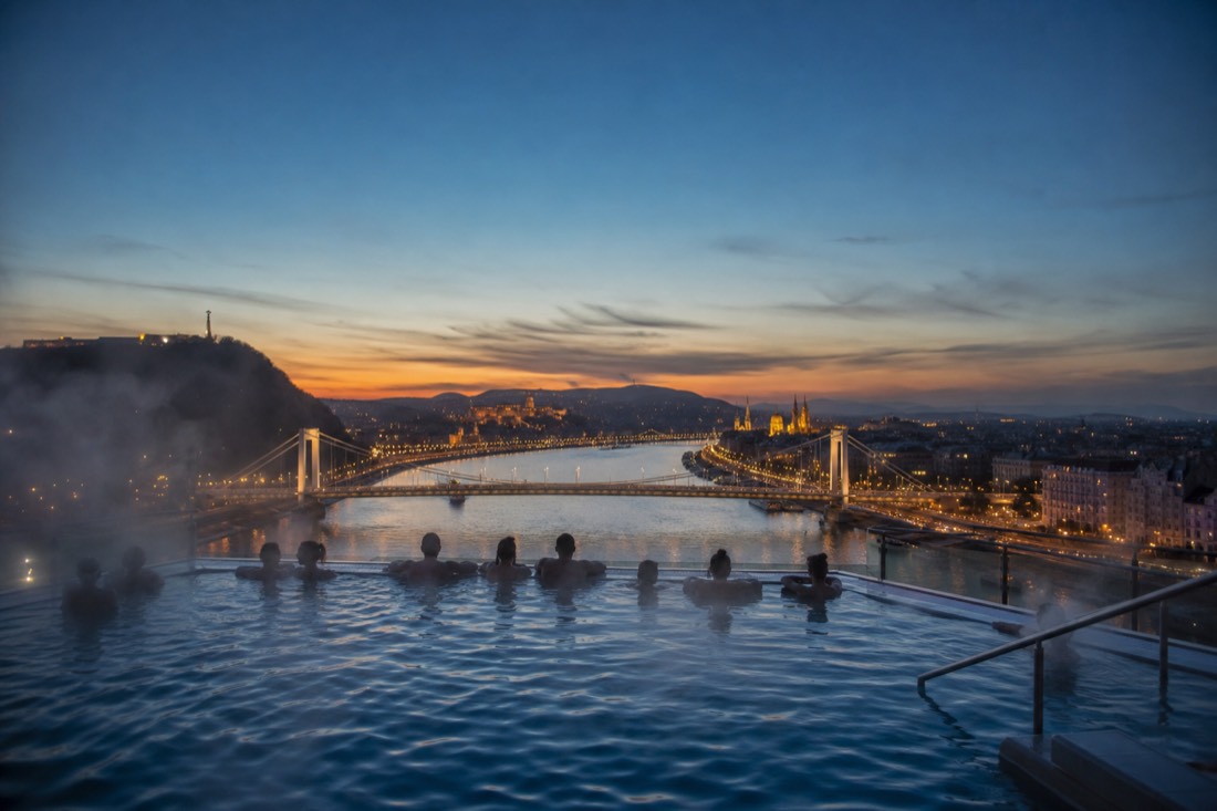 Steam rising from a rooftop thermal pool at dusk with the Danube and the lit Pest skyline beyond