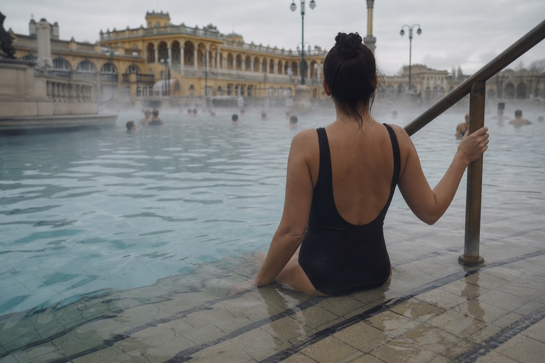 Adult sitting on the shallow steps of a mild outdoor thermal pool in Budapest with a handrail beside