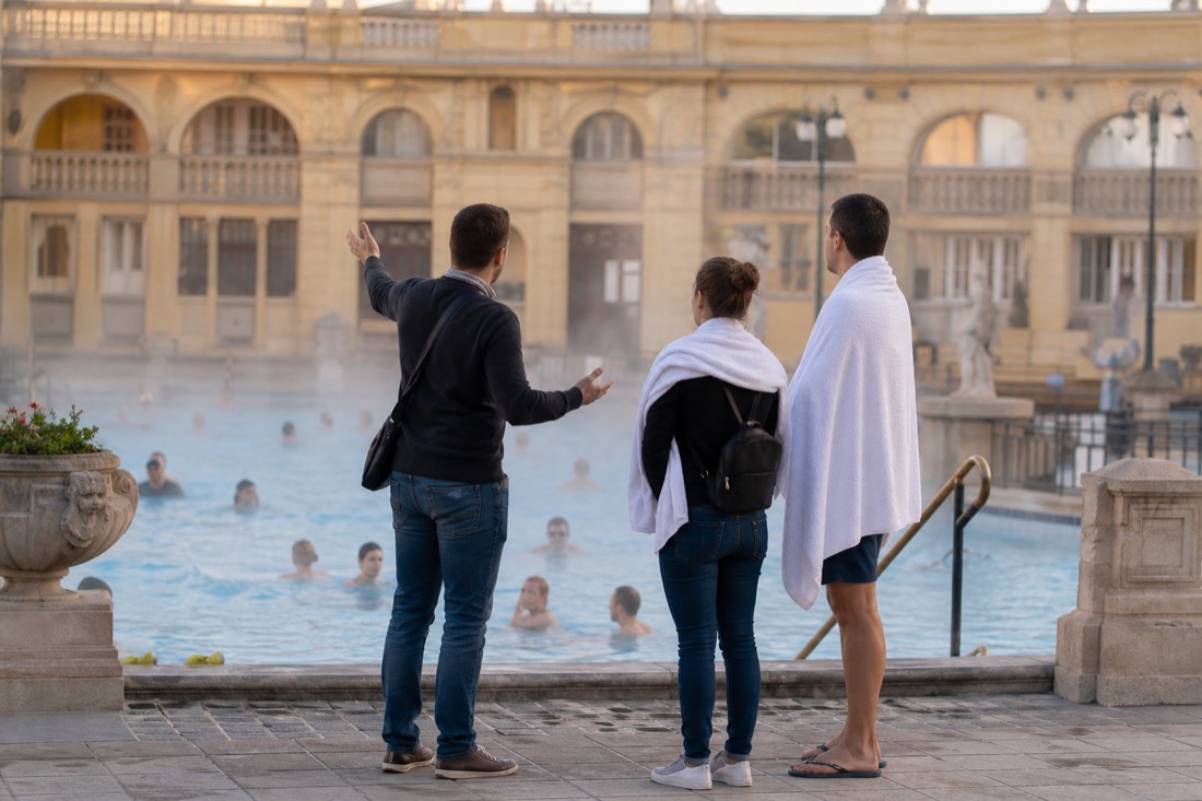 Guide gesturing toward a warm outdoor pool while speaking with two visitors at a Budapest bath courtyard