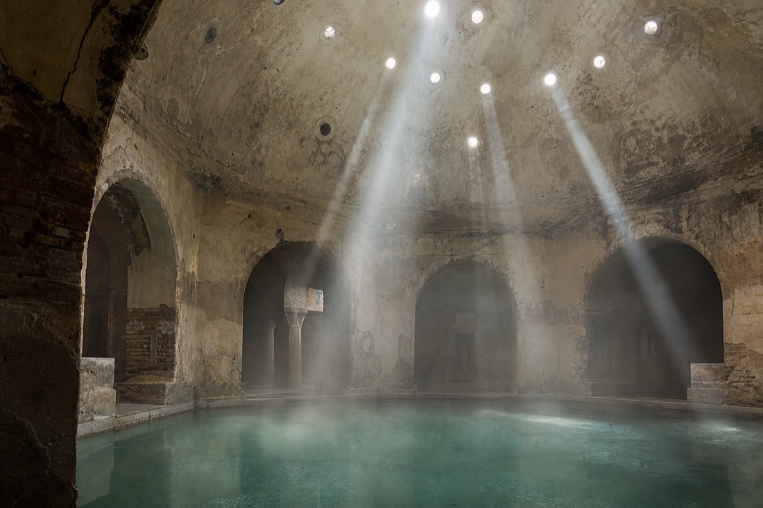 Shafts of morning light through small dome windows striking the steam above an old Ottoman bath pool in Budapest