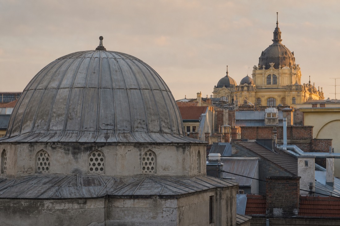 Aged stone dome of an Ottoman bath house against a Budapest skyline where a grand early twentieth century spa building rises behind it