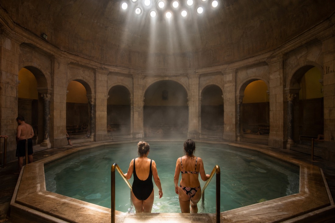 Two bathers seen from behind entering a warm domed pool through drifting steam in a historic Budapest Turkish bath